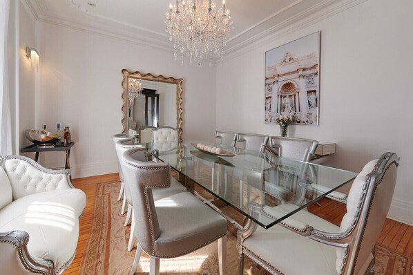 A dining room with a glass table, upholstered chairs, a chandelier, a large mirror, and a framed artwork on the wall.