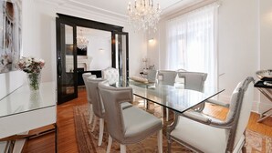 A dining area with a glass table, upholstered chairs, a chandelier, and a view of a living room through a doorway.