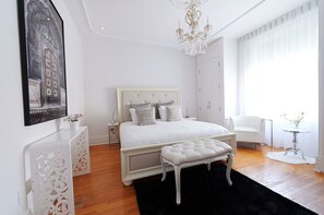 A bedroom with a bed, a chandelier, a white chair, and a small table with flowers.