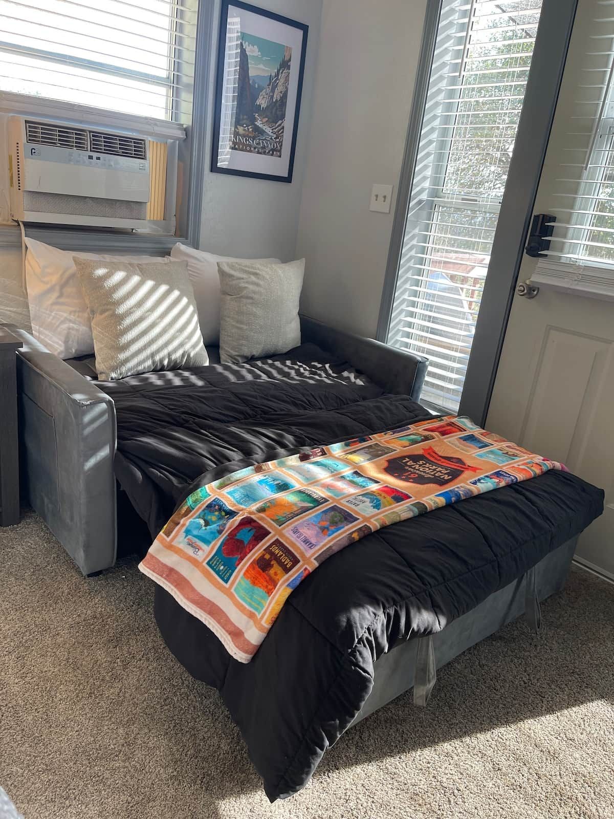 A single bed with a black mattress and a colorful quilt, a grey armchair, and a window with blinds.