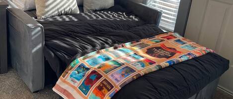 A single bed with a black mattress and a colorful quilt, a grey armchair, and a window with blinds.