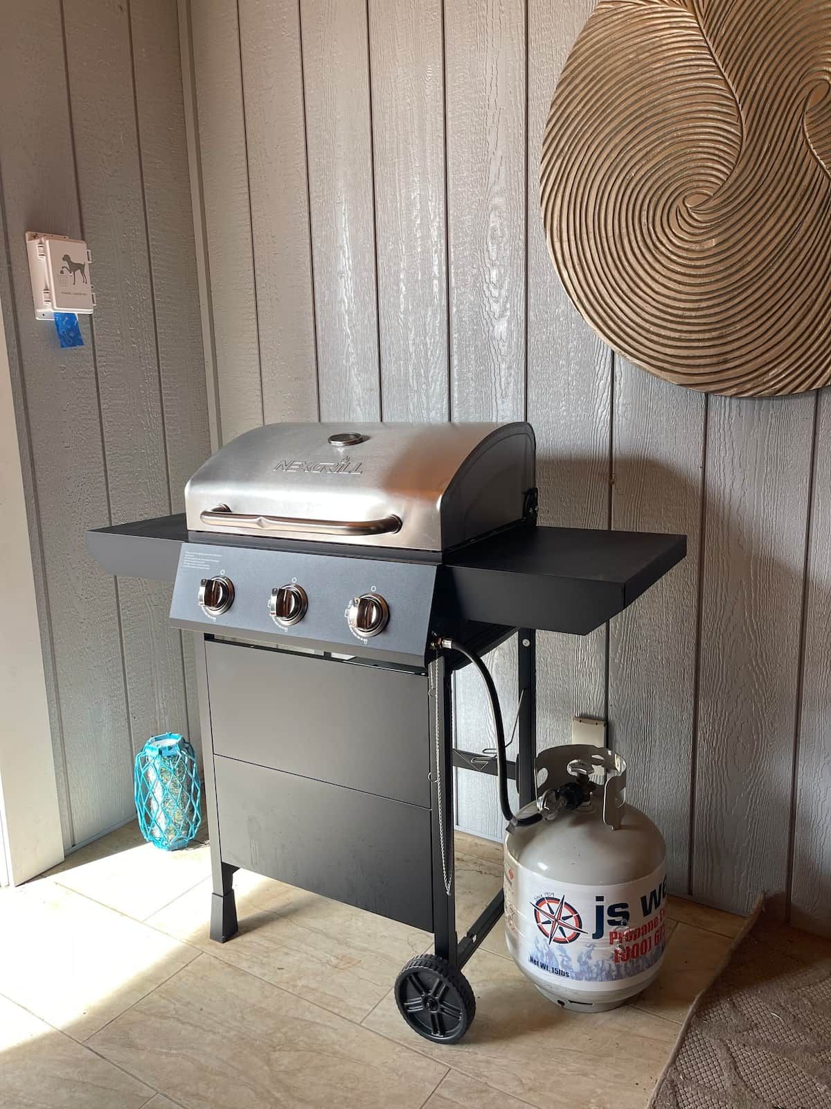 A portable grill with a gas tank on a wheeled cart, set against a wooden wall.