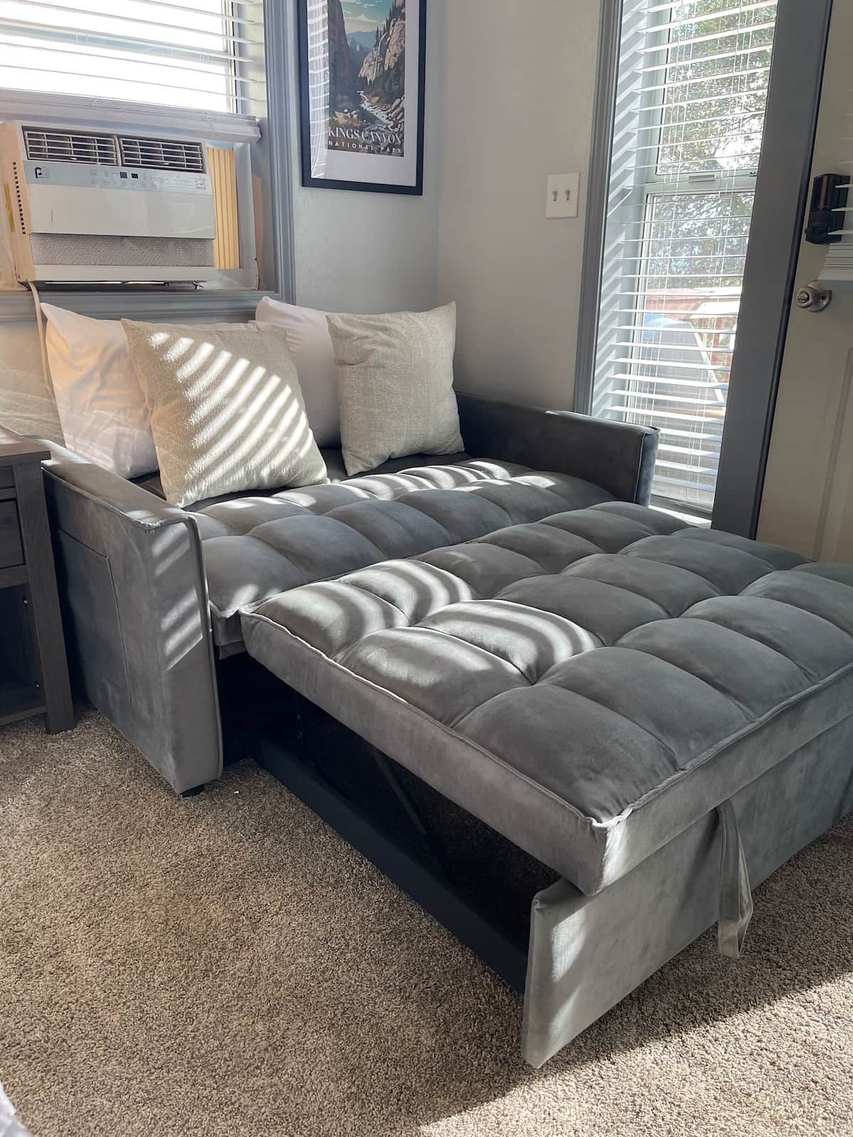 A modern bedroom with a grey upholstered bed, a nightstand, a framed picture on the wall, and a window with blinds.