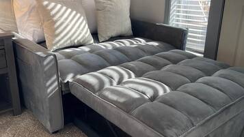 A modern bedroom with a grey upholstered bed, a nightstand, a framed picture on the wall, and a window with blinds.
