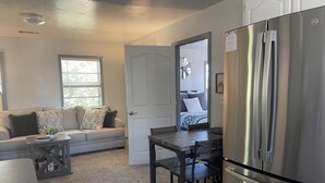 Un espacio habitable compacto con kitchenette, un sofá con cojines decorativos, una mesa de comedor y un refrigerador.