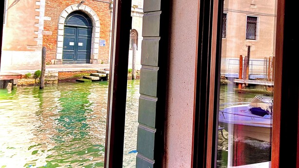A view through a window of a building with a brick facade and a blue door, overlooking a canal with a boat. - Canal View (Venezia)