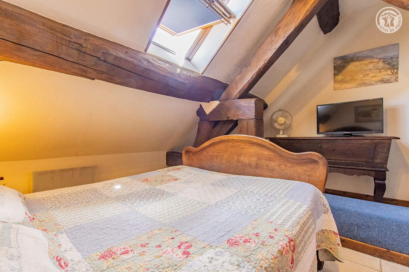 Une chambre avec un plafond en bois, un lit recouvert d’une courtepointe à motifs floraux, un bureau avec une télévision et un ventilateur.