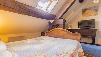 Une chambre avec un plafond en bois, un lit recouvert d’une courtepointe à motifs floraux, un bureau avec une télévision et un ventilateur.