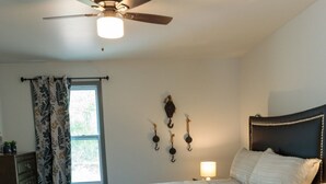 A bedroom with a bed, a ceiling fan, a window with curtains, wall-mounted hooks, a bedside lamp, and a small table with a plant.