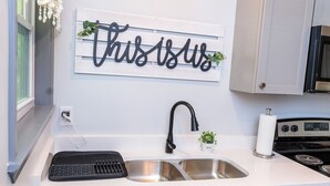 A kitchen with a double sink, a black keyboard, a small potted plant, and a wall-mounted sign that reads "this is us".