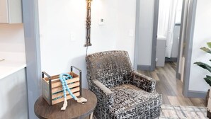A room with a patterned armchair, a small wooden table with a blue beaded rope, and a wall-mounted lamp.