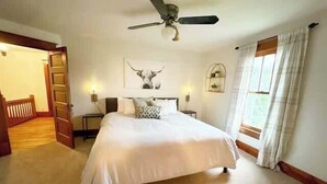 Un dormitorio con una cama grande, un ventilador de techo, una ventana con cortinas y una puerta que da a otra habitación.