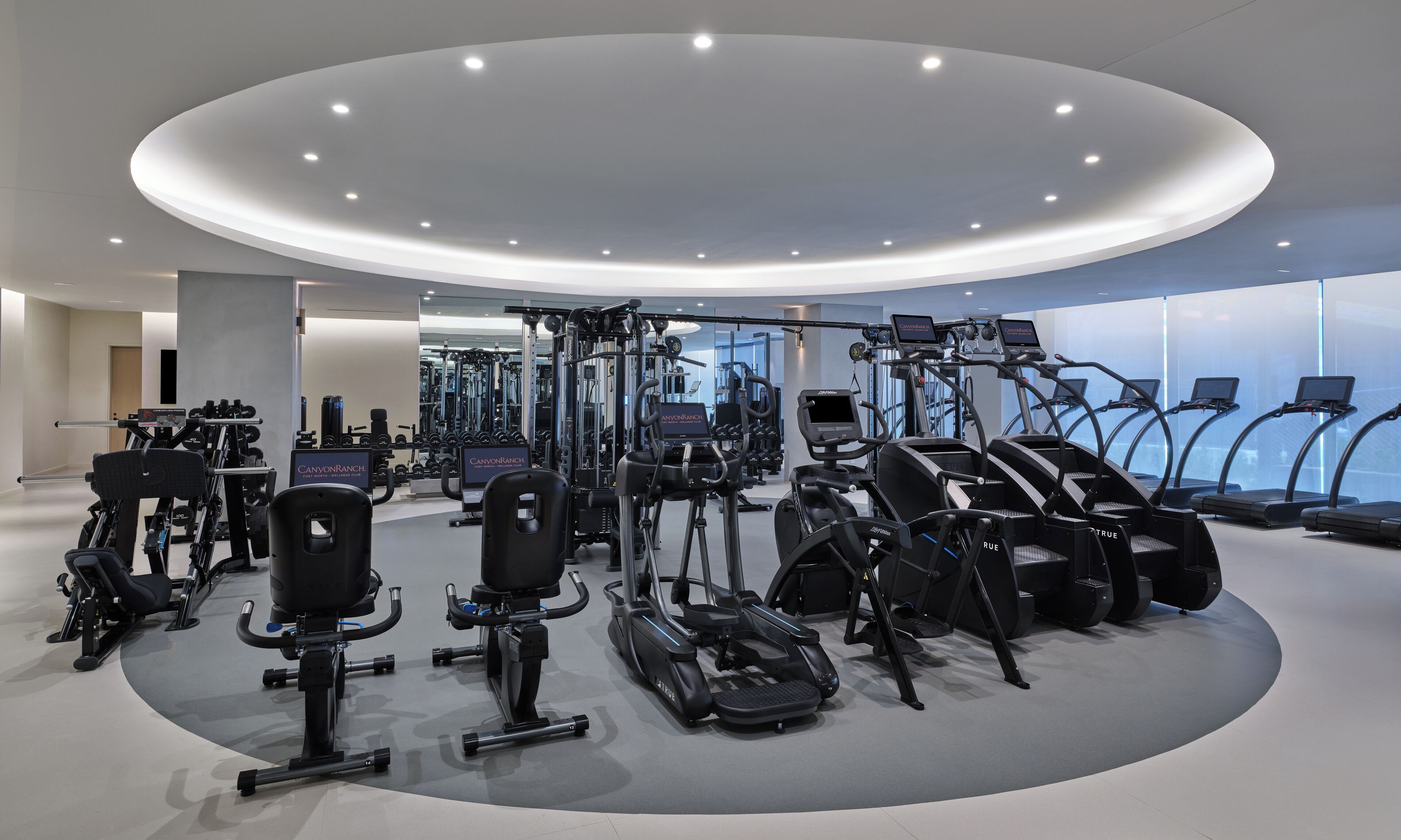 A modern gym with various cardio and strength training equipment, including treadmills and weight machines, arranged in a circular layout.