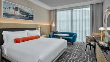 A hotel room with a large bed, a desk with a chair, a small round table, and a large window with curtains.