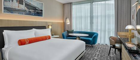 Une chambre d’hôtel avec un grand lit, un bureau avec une chaise, une petite table ronde et une grande fenêtre avec des rideaux.