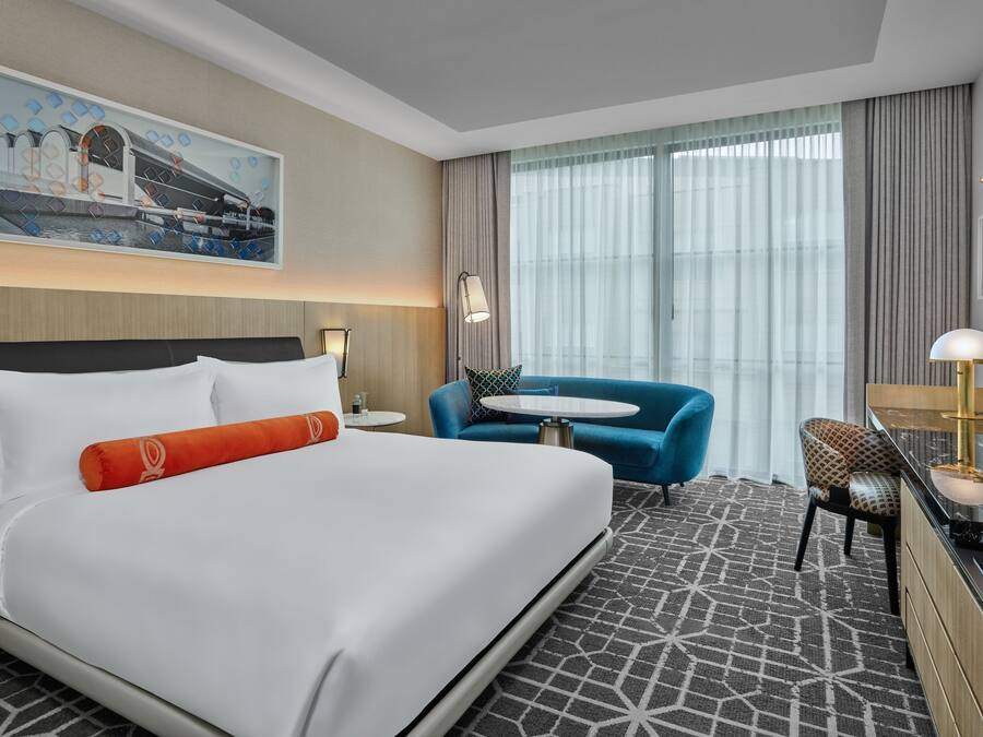 Une chambre d’hôtel avec un grand lit, un bureau avec une chaise, une petite table ronde et une grande fenêtre avec des rideaux.
