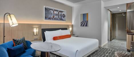 A hotel room with a bed, a round table, a blue armchair, and a wall-mounted artwork.