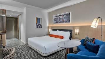 A hotel room with a bed, a blue armchair, a small round table, and a painting on the wall.