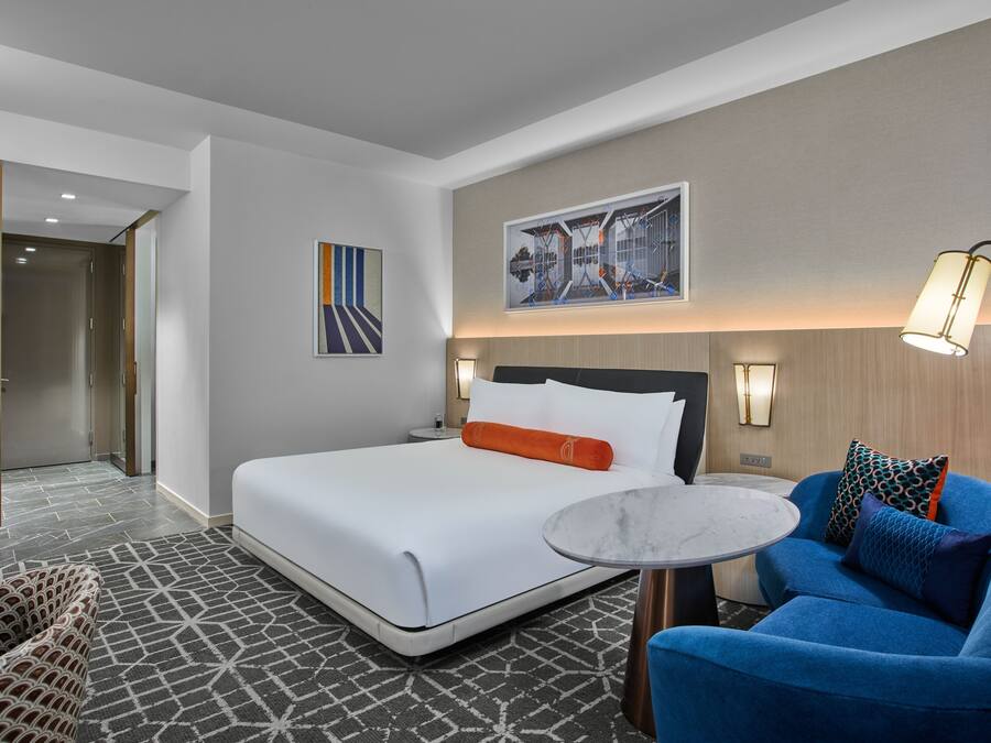 A hotel room with a bed, a blue armchair, a small round table, and a painting on the wall.