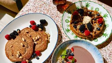 A table with three plates of food: pancakes with berries and nuts, waffles with berries and cream, and a bowl of chocolate with berries and granola.