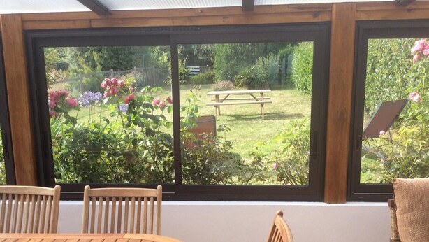 Un invernadero con una mesa y sillas de madera, vista a un jardín con flores y una mesa de picnic.