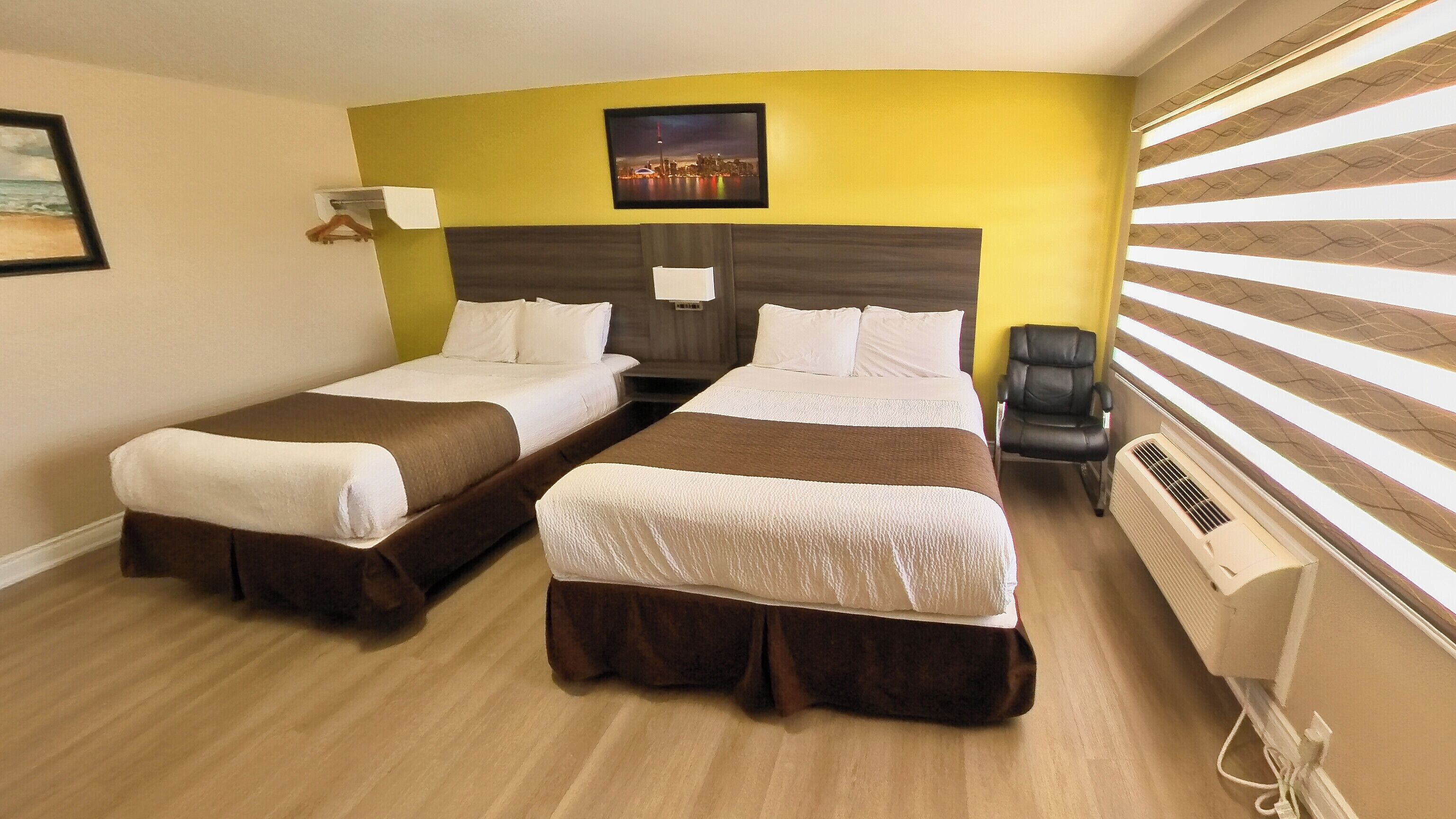 A hotel room with two beds, a cityscape picture on the wall, a chair, and a window with blinds.