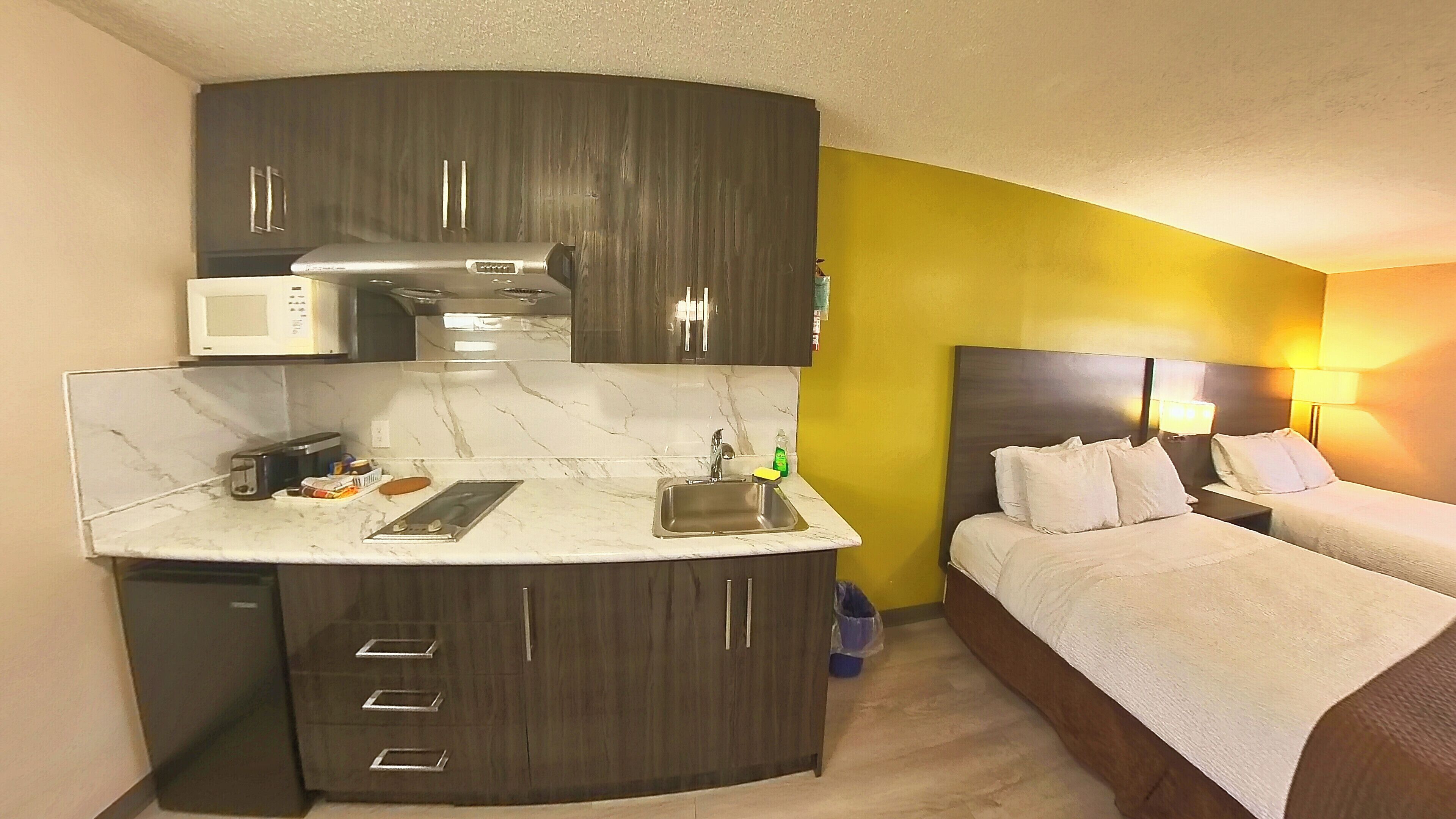 Une chambre d’hôtel compacte, dotée d’une kitchenette, d’un lit avec une literie blanche et d’une table de chevet avec une lampe.