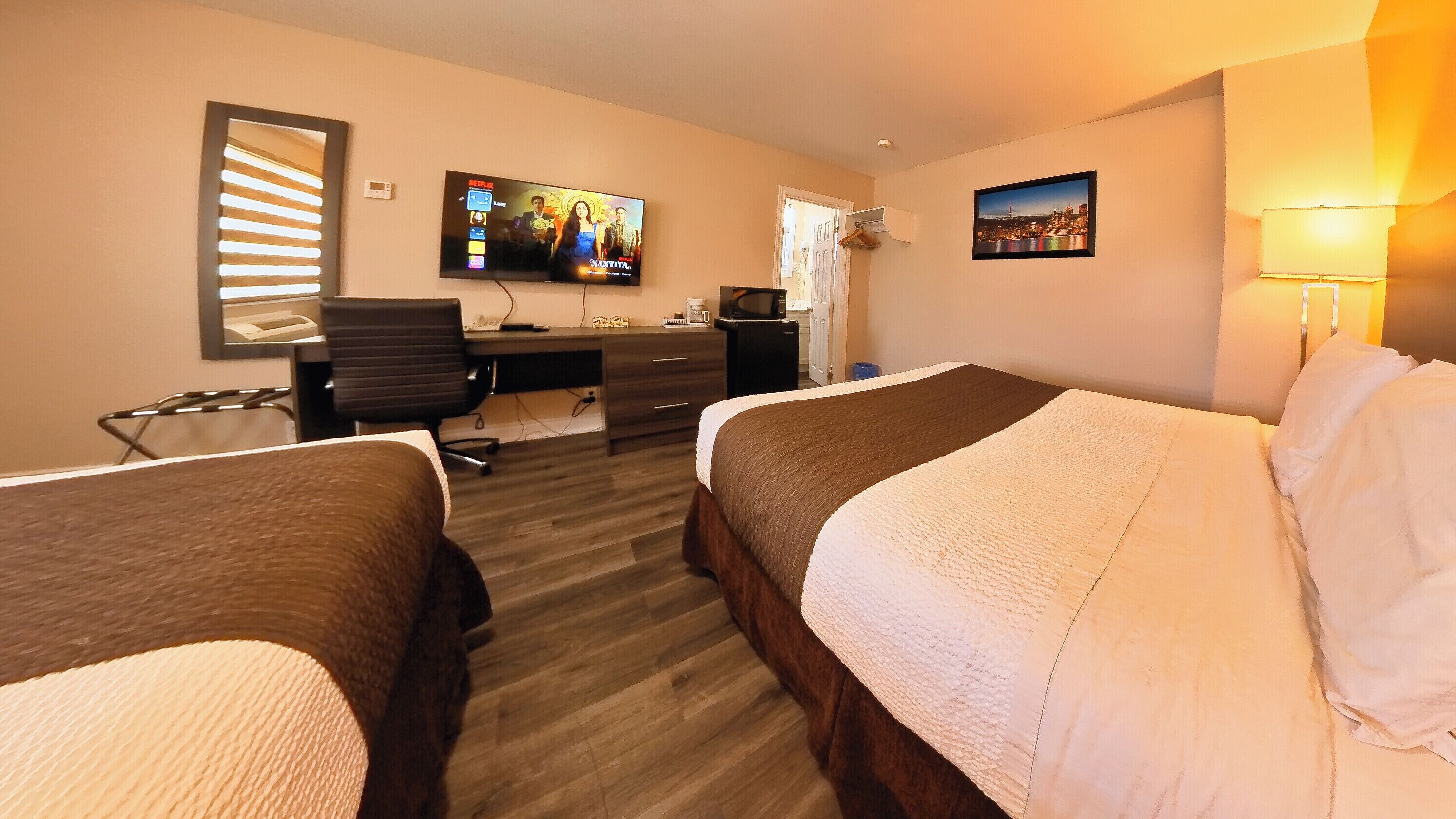 A hotel room with two beds, a desk, a TV, and a cityscape painting.