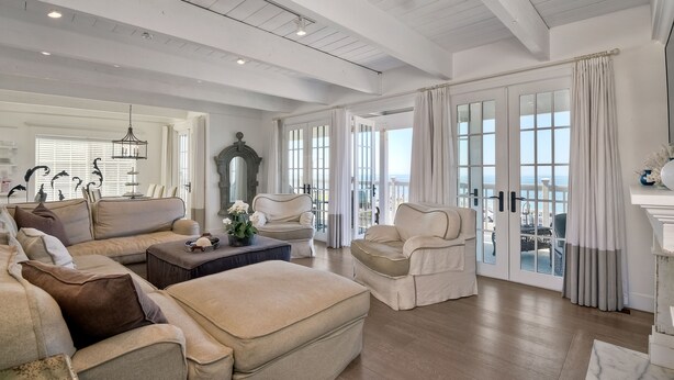 Un salón amplio con un sofá seccional grande, una mesa de centro y una chimenea. Hay ventanas grandes con cortinas y vistas al exterior.