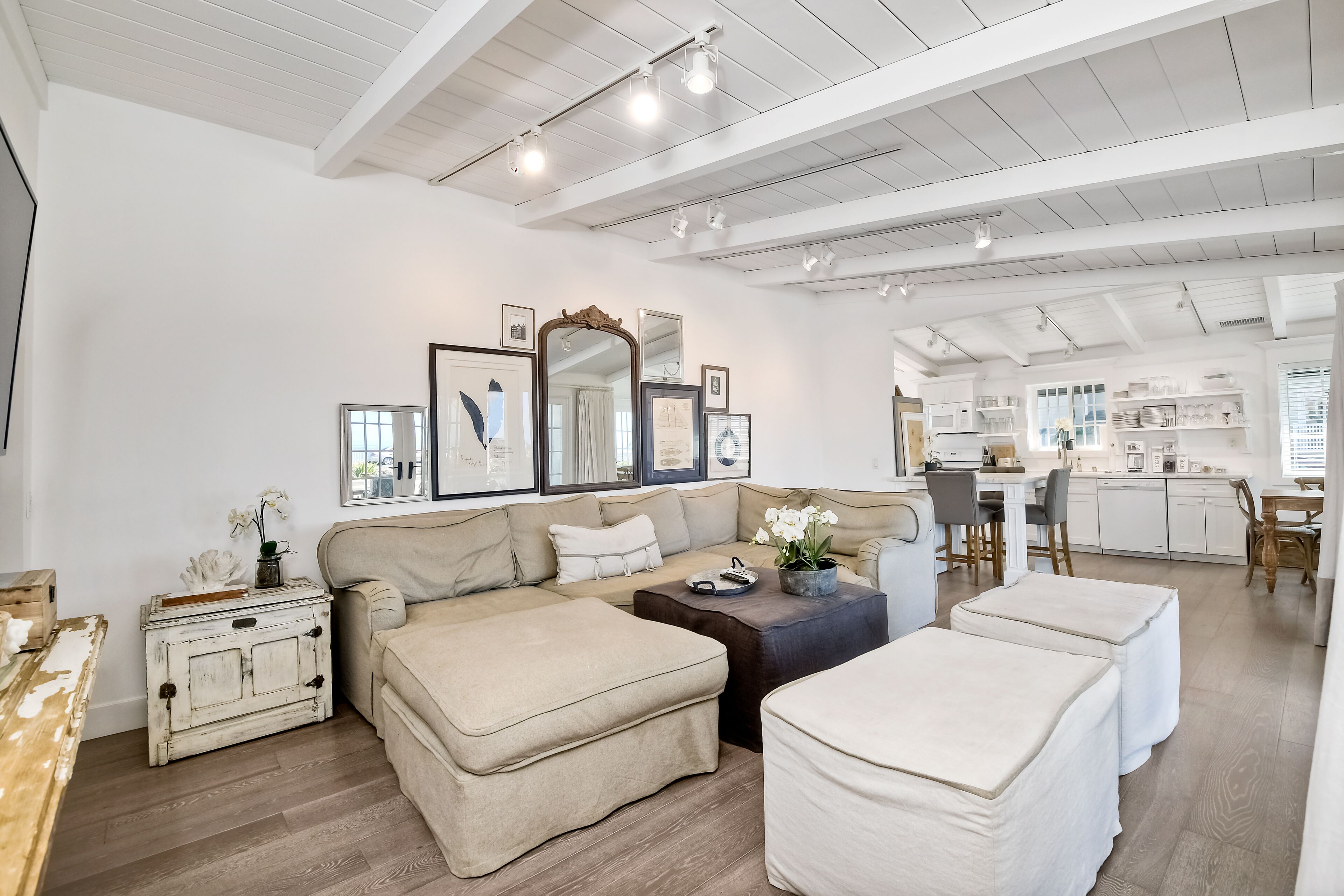 Un vaste espace de vie comprenant un canapé beige, des poufs, une table basse et une petite table d’appoint au style rustique. On aperçoit une cuisine au fond.