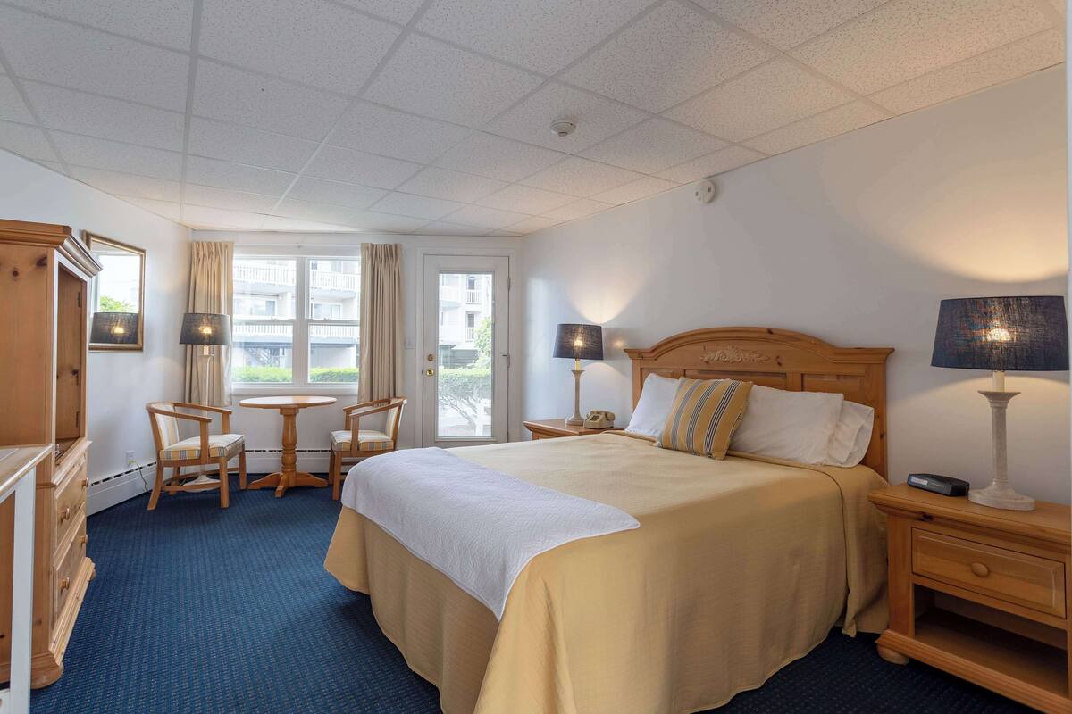 A hotel room with a large bed, a desk with a chair, a small table, and a nightstand with a lamp.