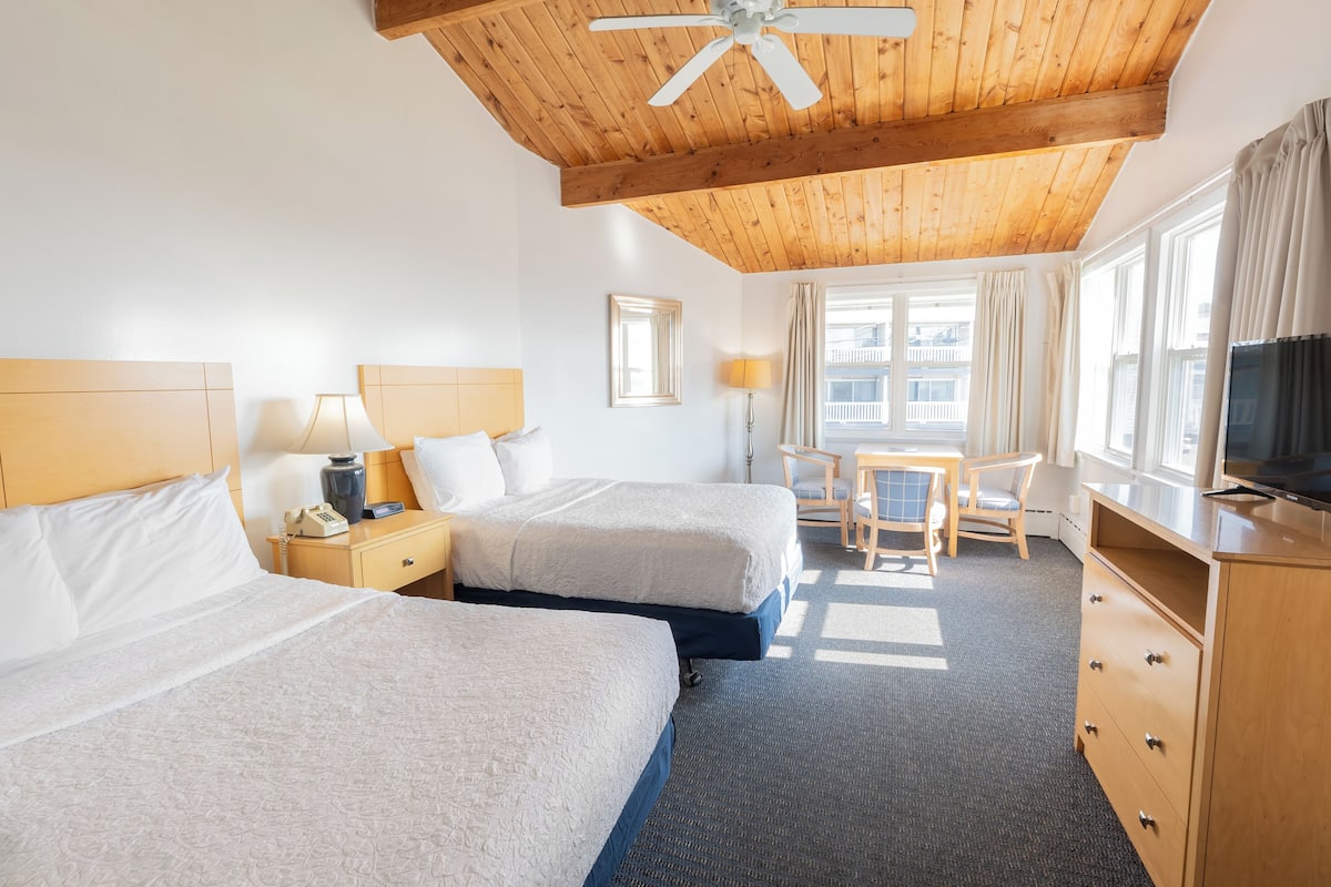 A hotel room with two beds, a desk, a chair, a television, and a ceiling fan.