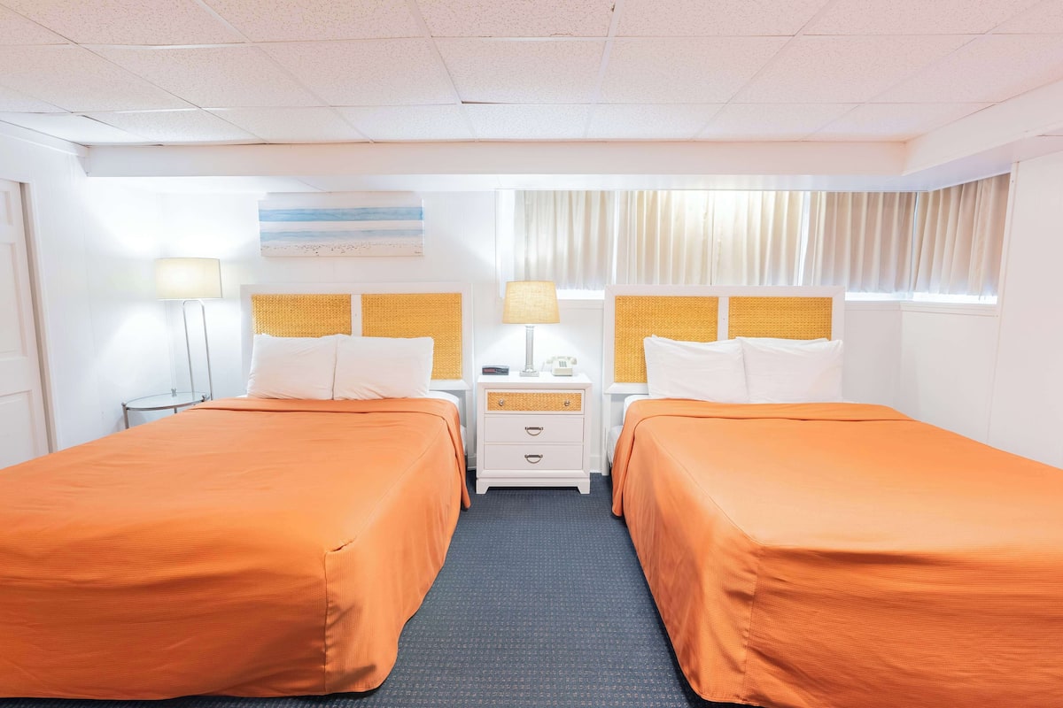A hotel room with two beds, a nightstand, a lamp, and a window with curtains.