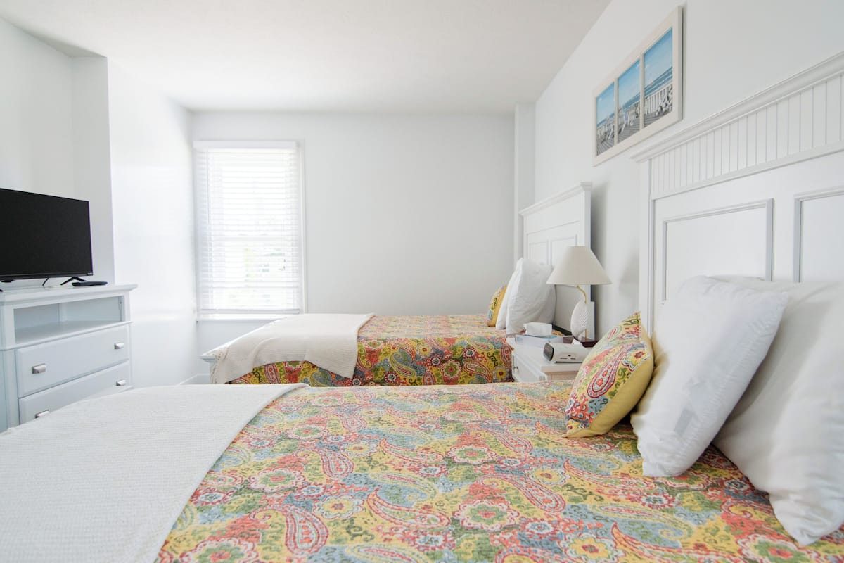 A bedroom with two beds, a television, a dresser, and a window with blinds.