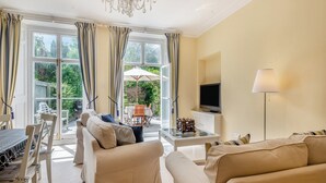 A spacious living room with a large sofa, a television, and a chandelier. There is a dining area with a table and chairs, and a view of the garden through large windows. - 3 bedroom accommodation in Cromer (Cromer)