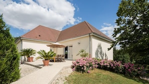 Ein zweigeschossiges Haus mit rotem Ziegeldach, einer Terrasse mit Tisch und Stühlen und einem Garten mit rosa Blumen.