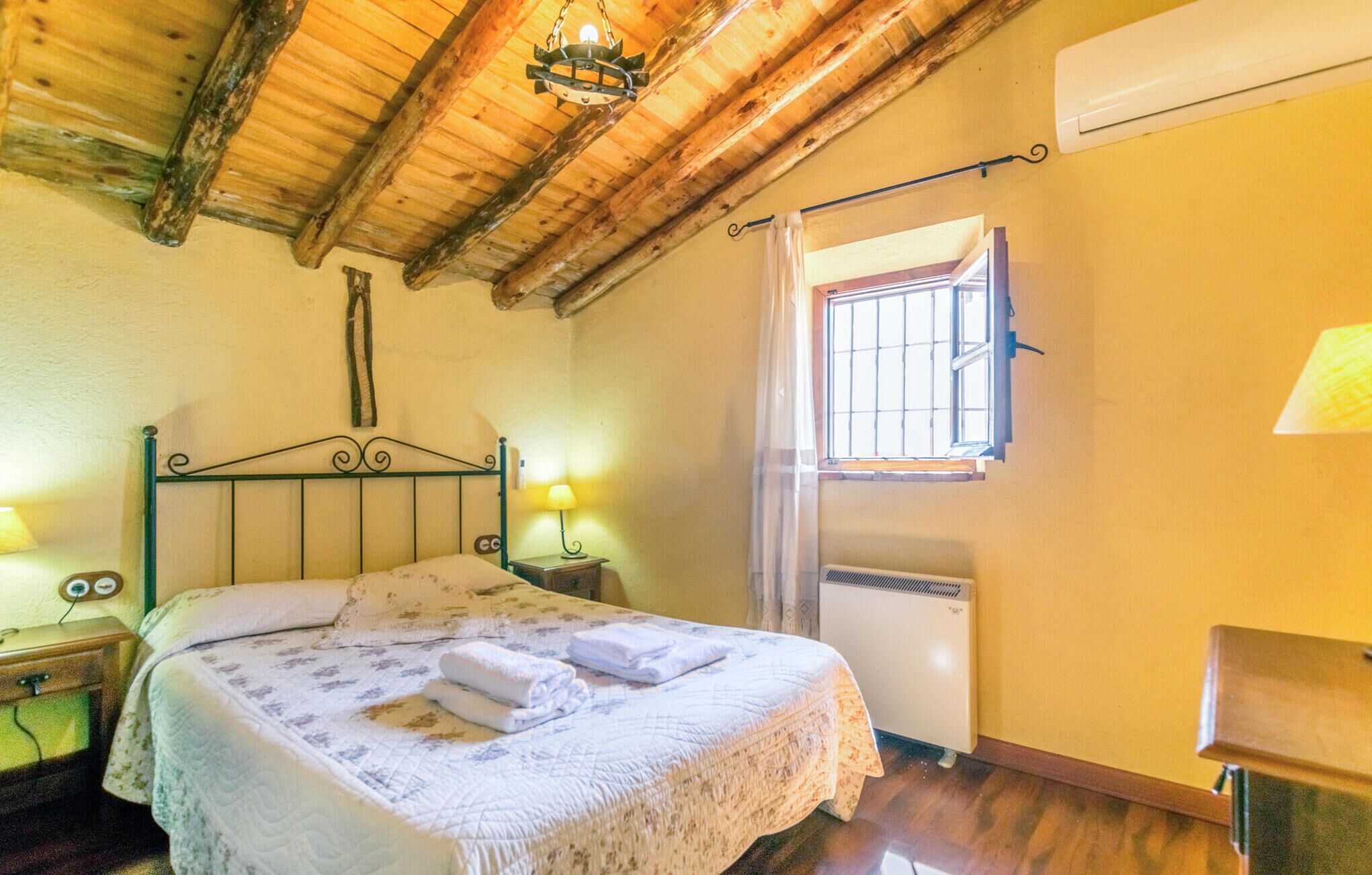 Een slaapkamer met een houten plafond, een bed met een witte sprei, twee bedlampen, een raam met tralies en een airconditioning.