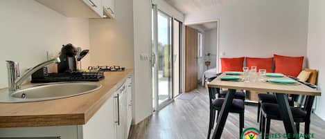 A modern kitchen with a wooden countertop, stainless steel sink, and a coffee machine. There is a dining area with a table set for four, featuring black chairs and a red cushion.