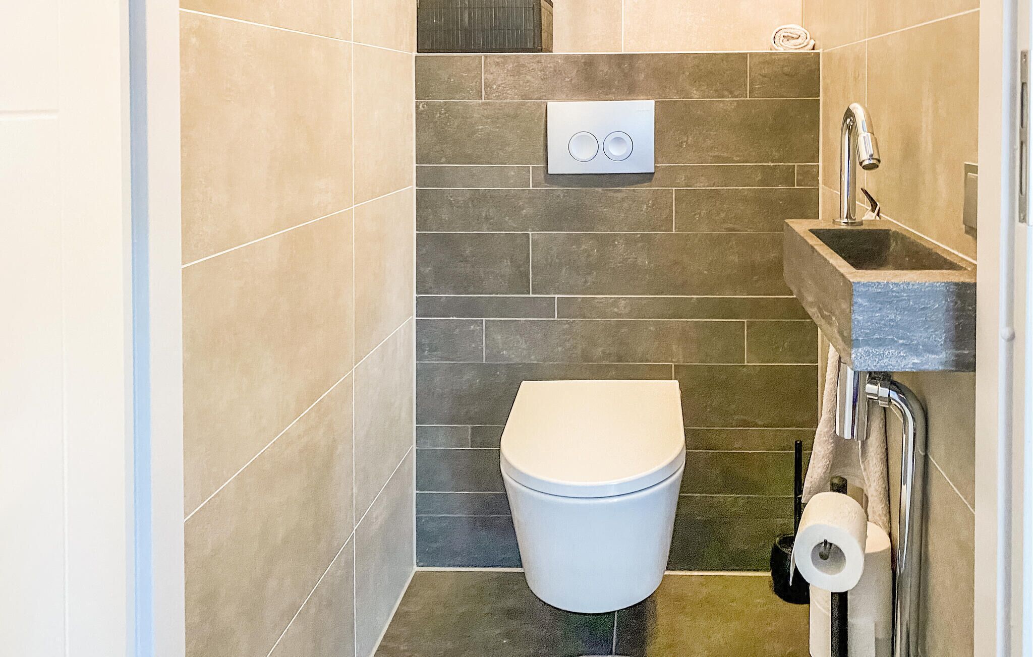 Un baño pequeño con inodoro, un mecanismo de descarga en la pared, un lavamanos con grifo y un dispensador de papel higiénico.