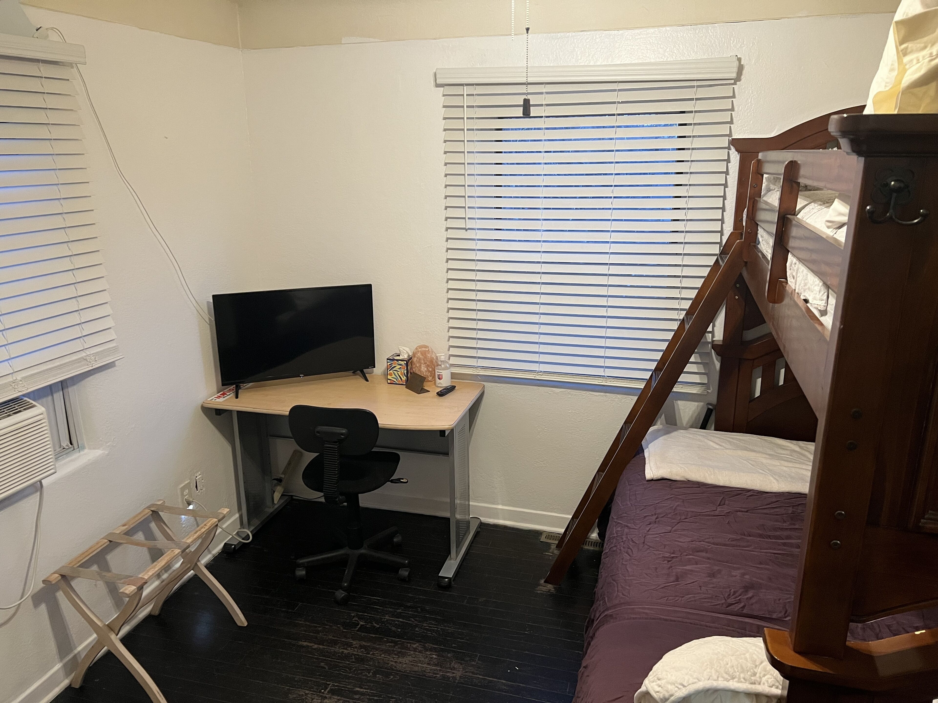 Ein Schlafzimmer mit einem Bett, einem Schreibtisch mit Computer und einem Fenster mit Jalousien.