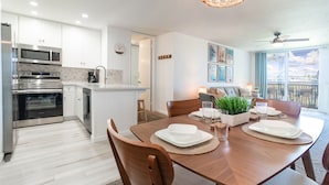 A modern kitchen with stainless steel appliances, a dining area with wooden furniture, and a view of a cityscape through large windows. - Sunset Harbor and Ocean Views, Modern Coastal Design, Beachfront Resort (Oceanside)