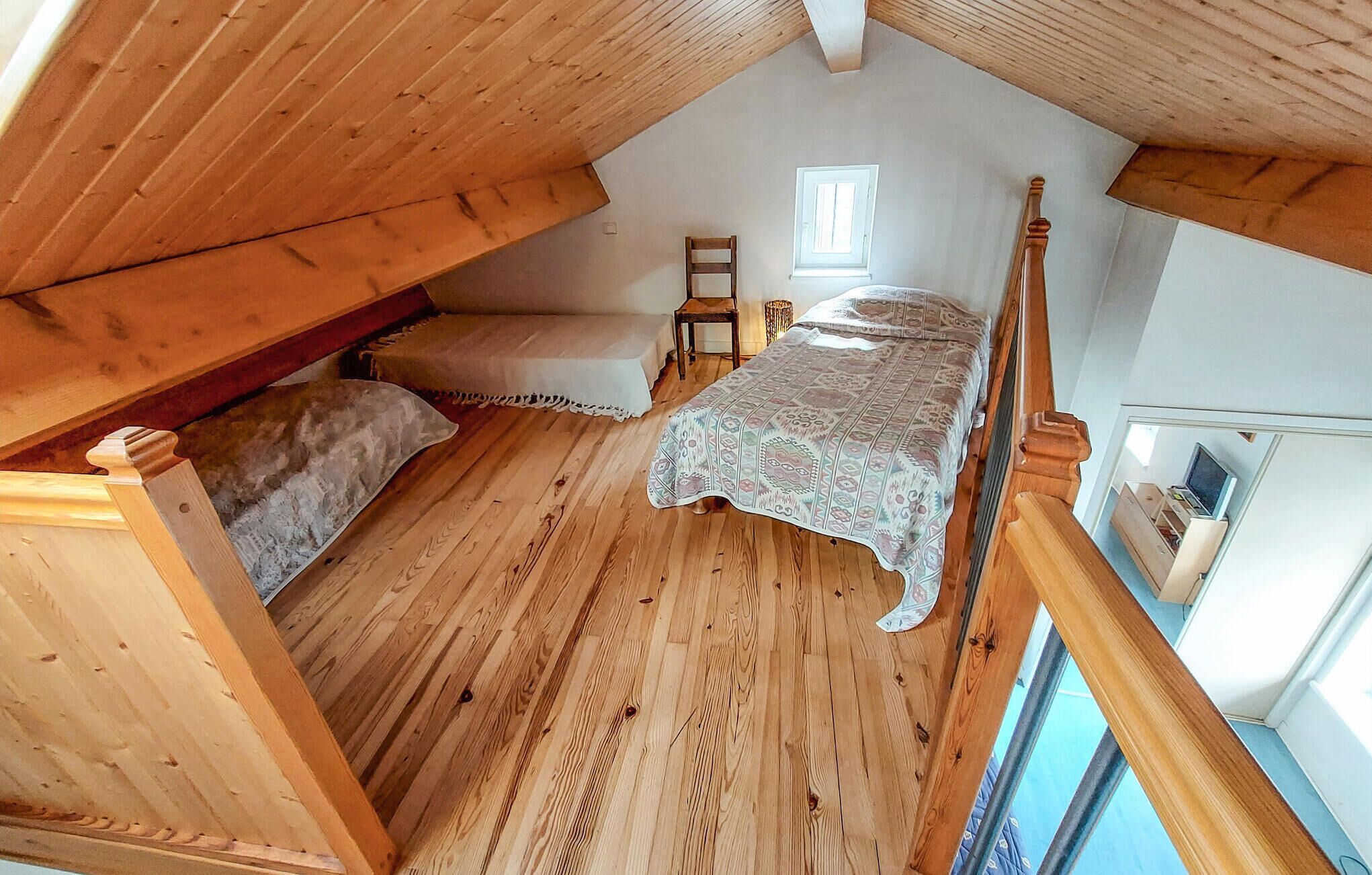 A bedroom with a wooden floor, a bed with a patterned bedspread, a chair, and a window.