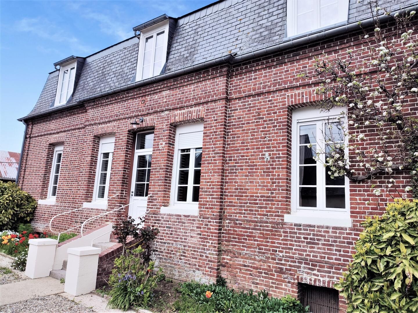 A two-story brick building with white-framed windows, a visible chimney, and a small garden area with steps leading to the entrance.