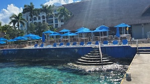 A poolside area with blue umbrellas and lounge chairs, a staircase leading into the water, and a building with a thatched roof in the background. - Luxury Condo Steps from Ocean with Great Views at The Landmark Resort of Cozumel (San Miguel de Cozumel)