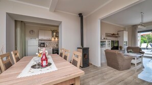 Uma sala de jantar com mesa e cadeiras de madeira, uma cozinha com eletrodomésticos modernos, uma lareira a lenha preta e uma área de estar com móveis de vime.