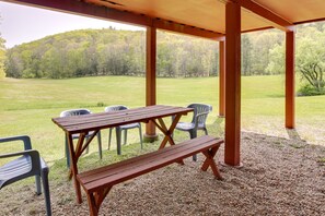 Een overdekte picknickplaats met een houten tafel en banken, omringd door plastic stoelen, met uitzicht op een grasveld en een bebost heuvel.