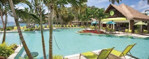 Un espace piscine aménagé dans un complexe hôtelier, avec des chaises longues, des palmiers et un bâtiment à toit de chaume.
