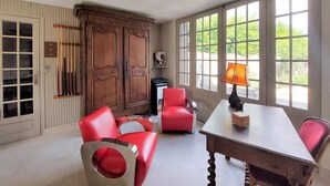 Ein Zimmer mit einem Holzkabinett, roten Stühlen, einem Tisch und großen Fenstern mit Blick auf Grünflächen.