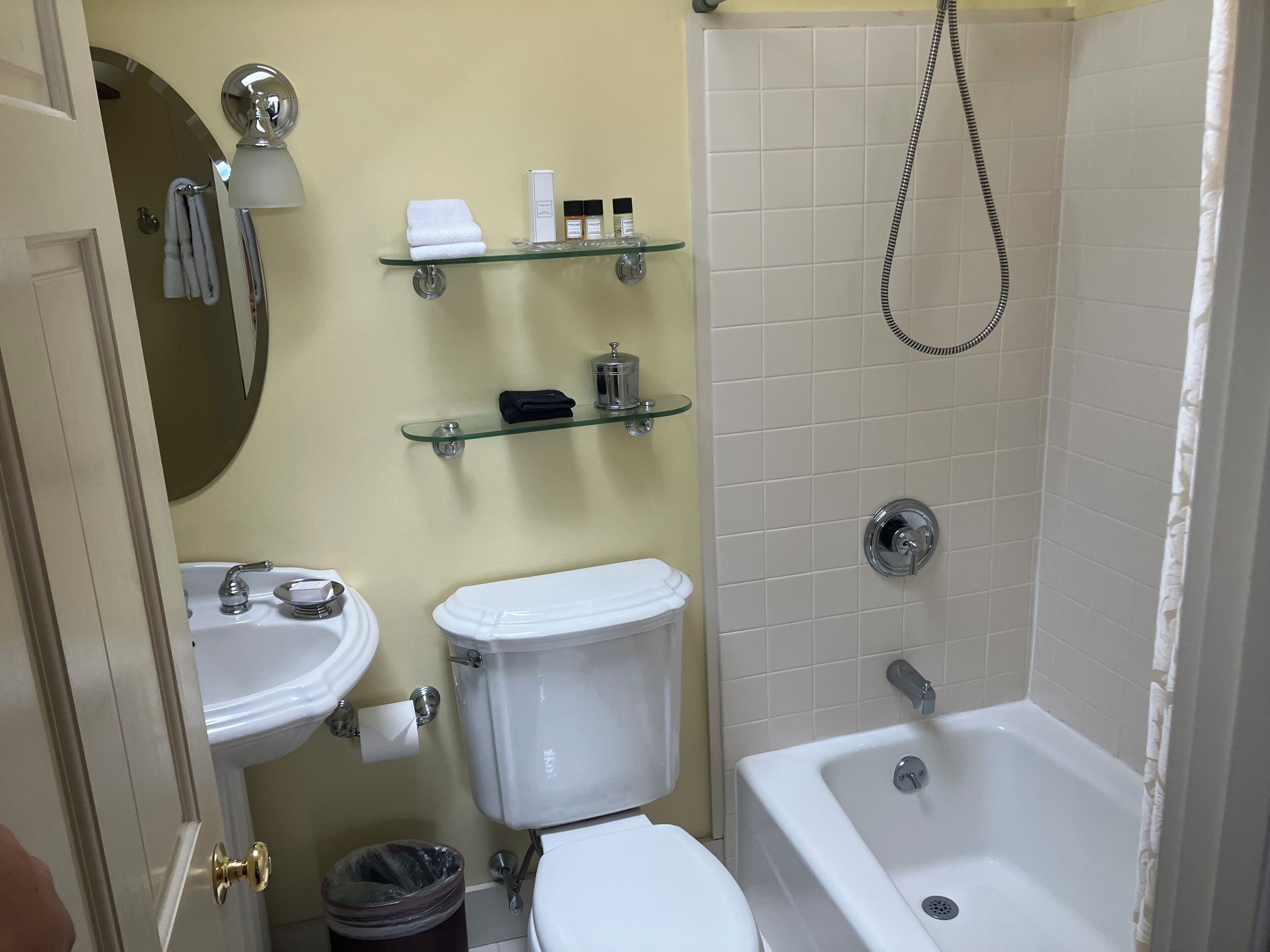 Um banheiro com vaso sanitário, pia e banheira. Há prateleiras de vidro acima do vaso sanitário, com produtos de higiene pessoal.