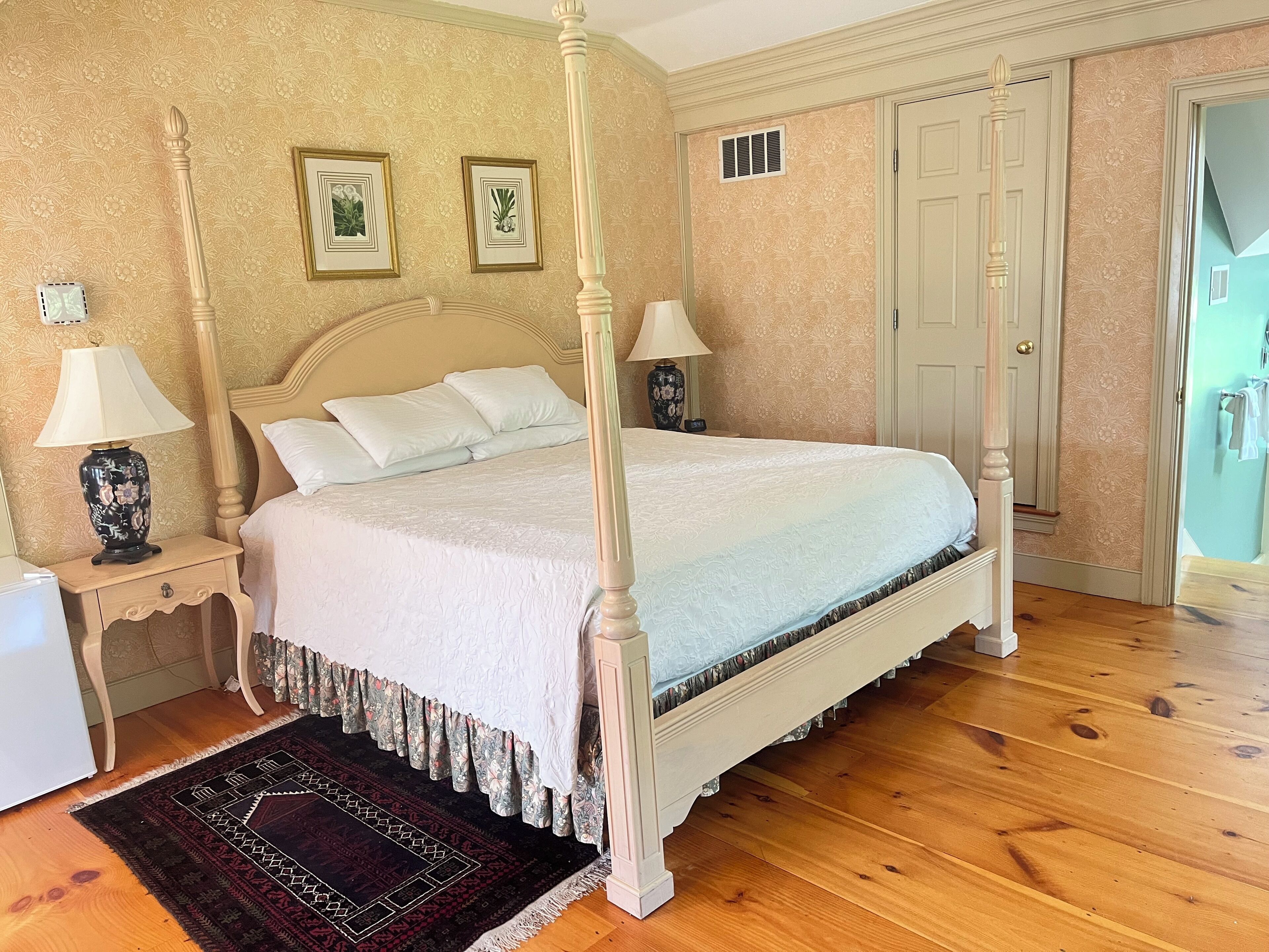 Un lit à baldaquin avec une couvre-lit blanche, deux tables de chevet avec des lampes, un tapis à motifs et une porte ornée d’un panneau décoratif.